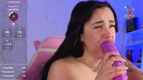 Media: Video of a young woman with long black hair, light skin, and a slender physique, crying while performing oral sex with a large purple dildo. Background shows a gaming setup with various game elements, including a pink gaming chair.