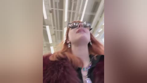 Media: Video of a fair-skinned woman with long red hair, wearing reflective sunglasses and a maroon fur coat, adjusting her hair in a modern, industrial setting with white ceilings and fluorescent lights.