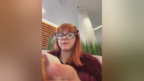 Media: Video of a young woman with fair skin, red hair, and glasses, wearing a maroon fur coat, reading a book in a modern room with green plants and wooden blinds.