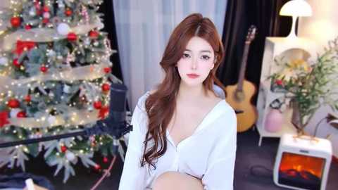 Media: Video of a fair-skinned, slender Asian woman with long, wavy brown hair, wearing a white cardigan, standing in a cozy, decorated living room with a Christmas tree, a fireplace, and guitar.