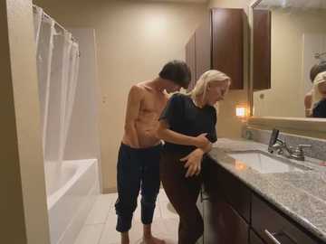 Media: Video of a shirtless man with medium build and dark hair, leaning over a blonde woman in a bathroom.