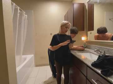 Media: A video of a blonde woman in a black dress and man in a white tank top using a smartphone in a bathroom with beige walls and a granite countertop.