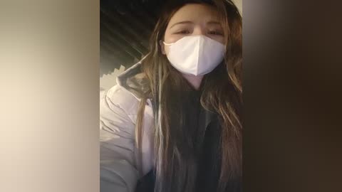 Media: Video of a young Asian woman with long, straight brown hair wearing a white face mask, a grey hoodie, and a white t-shirt. She appears to be indoors, possibly in a dimly lit room with blurred background.