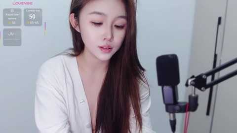 Media: Video of an East Asian woman with long brown hair, light skin, wearing a white cardigan, singing into a microphone, against a plain white background.