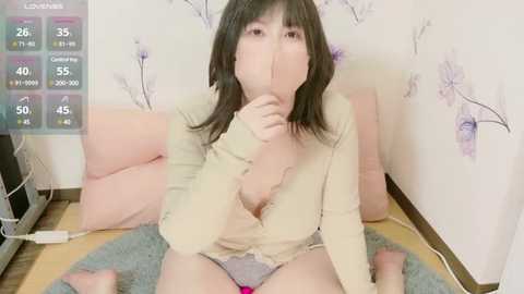 Media: Video of a light-skinned woman with straight black hair, wearing a beige shirt and grey panties, sitting on a blue rug, partially covering her face with her hand, in a room with floral wallpaper and electronic device icons displayed.