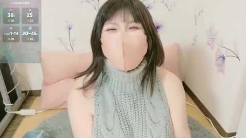 Media: Video of an Asian woman with straight black hair, wearing a gray knitted turtleneck sweater, and a gray face mask, sitting on a pink couch with floral wallpaper in the background.