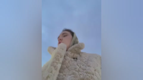 Media: Video of a woman with light skin and dark hair, wearing a fluffy, cream-colored hat and coat, gazing upward with a thoughtful expression. The background is a soft, cloudy sky.