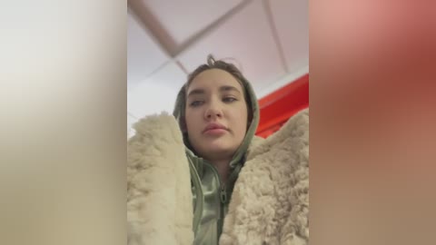 Media: Video of a young woman with light skin and dark hair, wearing a green hooded jacket and a fluffy cream coat, indoors with a red wall in the background.