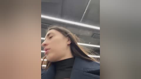 Media: Video of a young woman with long brown hair, wearing a black turtleneck and navy blazer, captured in a dimly lit, modern room with fluorescent lighting and exposed ceiling panels.