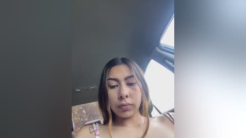 Video of a Latina woman with long, straight, dark brown hair and light brown skin. She wears a light pink, off-shoulder top, sitting in a car with a dark gray interior.