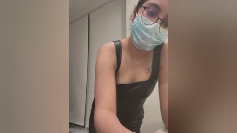 Media: Video of a young woman with light skin and glasses, wearing a black leather dress and a blue surgical mask, standing in a minimalistic room with white walls and closed closet doors.