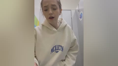 Media: Video of a young woman with fair skin, brown hair in a ponytail, wearing a beige hooded sweatshirt with \"WILLIAMSON\" logo, standing in a narrow, white-tiled hallway.