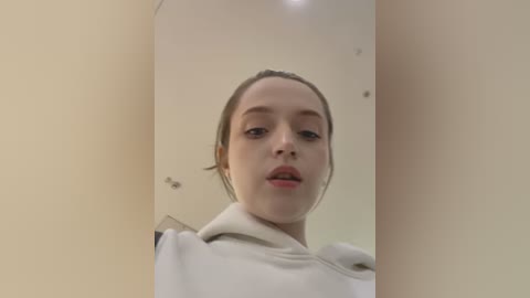 Media: A video of a young woman with fair skin, brown hair tied back, wearing a beige hoodie, standing in a beige room with a ceiling light fixture.