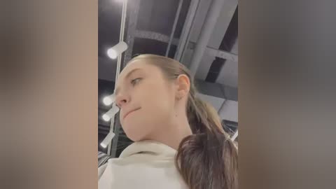 Media: Video of a young woman with light skin and long brown hair tied back, wearing a white top, looking upward. The background features industrial pipes and lights, suggesting an indoor, possibly urban setting.