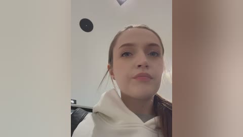 Media: Video of a young woman with fair skin and light brown hair, wearing a white hoodie, standing in front of a black chair and white wall with a ceiling light.