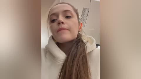 Media: Video of a fair-skinned woman with long brown hair tied in a ponytail, wearing a cream-colored hoodie, looking down slightly with a neutral expression in a beige hallway.