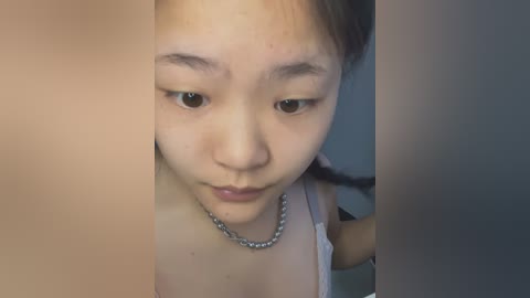 Media: A close-up video of a young Asian girl with light skin and straight black hair, wearing a white tank top and a silver chain necklace. She has a neutral expression and acne on her cheeks.