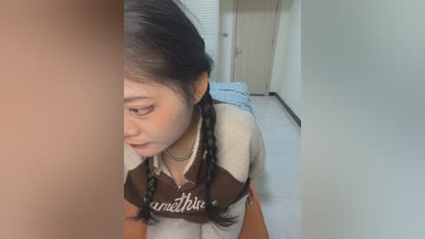 Media: Video of a young Asian woman with long black hair in braids, wearing a brown and beige striped sweater, looking sad in a minimalist bedroom with white walls and a blue bedspread.