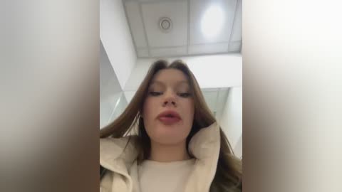 Media: Video of a Caucasian woman with straight brown hair, wearing a beige coat, standing in a bright, modern office with white walls and a circular light fixture.