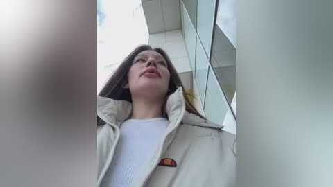 Media: A video of a young woman with fair skin and long brown hair, wearing a beige jacket with a red and yellow logo patch, standing outside a modern glass building.