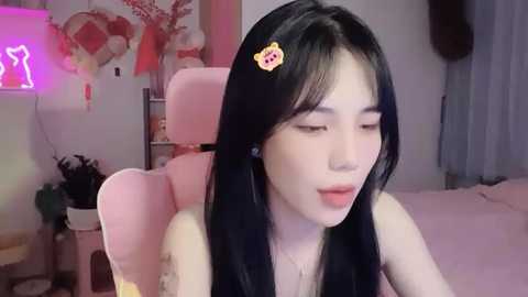 Media: Video of a young East Asian woman with long black hair, wearing a pink hair clip, sitting on a pink gaming chair in a cozy bedroom with pink and white decor.