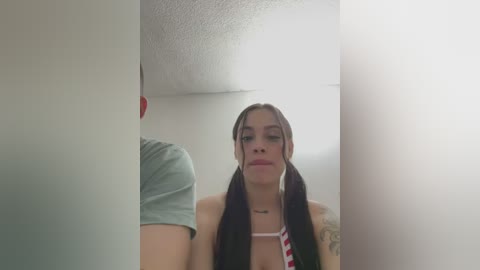 Media: Video of a young woman with long dark hair styled in pigtails, wearing a striped necktie, standing in a white room with a textured ceiling and partially visible person on the left.