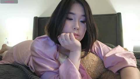 Media: Video of an Asian woman with shoulder-length black hair, lying on a bed in a pink satin robe, holding a teddy bear, in a softly lit bedroom.