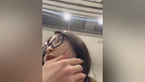 Media: Video of a young woman with glasses, dark hair, and light skin, touching her neck. She is indoors with a modern, industrial ceiling and fluorescent lights.