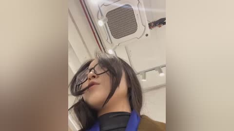 Media: Video of a woman with straight black hair, wearing glasses, leaning forward in a plane aisle, with beige walls and a white ventilation unit in the background.