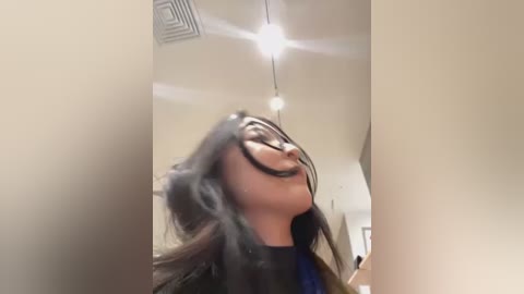 Media: A video of a woman with long brown hair, wearing black glasses, laughing with her head thrown back, taken indoors under a bright light fixture.