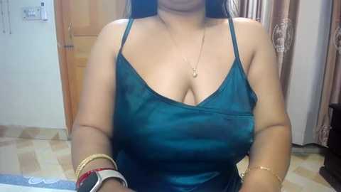 Media: Video of a woman with medium skin tone, wearing a teal satin camisole that reveals cleavage, multiple gold bracelets, and a gold necklace, seated in a room with wooden door and beige curtains.