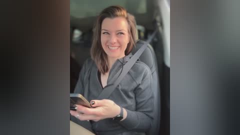 Media: A video shows a smiling woman in a car, wearing a gray hoodie, holding a smartphone, with a seatbelt across her chest. The background is blurry.