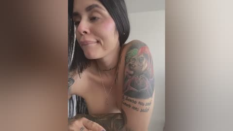 Media: Video of a smiling, tattooed, topless woman with shoulder-length black hair and fair skin, leaning forward in a bedroom with a plain white wall.