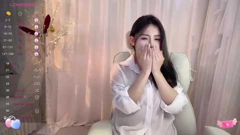 Media: Video of a young East Asian woman with long black hair, wearing a white shirt, covering her face with her hands, seated in a white chair against light pink curtains.