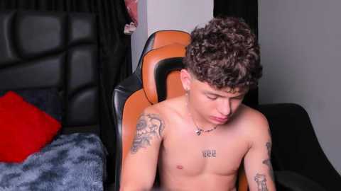 Video of a shirtless, curly-haired, tattooed young man with a lean physique, seated on a black leather gaming chair. He's wearing a necklace. Background includes a bed with blue sheets and a red pillow.