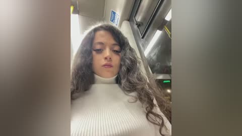 Media: A video of a woman with curly, dark hair wearing a white ribbed turtleneck sweater. She is in a subway car, with fluorescent lights and metal surfaces in the background.