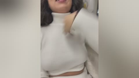 Media: A video of a woman with medium brown skin, shoulder-length dark hair, wearing a white turtleneck sweater, her cleavage partially visible. She stands in a dimly lit room, partially blurred background.