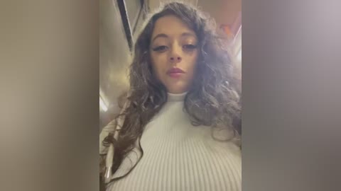 Media: Video of a young woman with long, wavy, dark hair, wearing a white ribbed turtleneck sweater, posing in an elevator. Her skin tone is light, and she has a neutral expression.