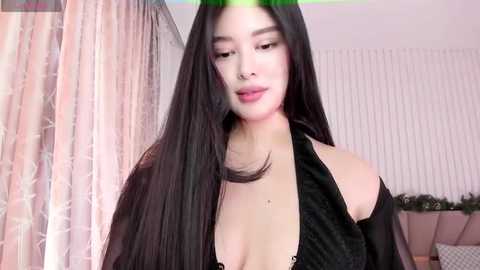 Media: Video of a young Asian woman with long black hair, light skin, and a slender figure. She wears a black halter top, revealing cleavage, in a softly lit room with pink curtains and white vertical blinds.