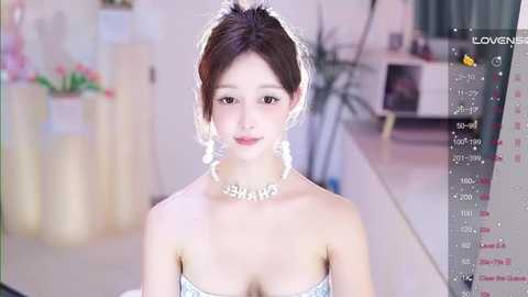 Media: A video of a young Asian woman with fair skin, brown hair styled in an updo, wearing a strapless white dress and a necklace with the word \"JENN\" in gold letters. The background is a blurred indoor setting with a potted plant and a white wall.