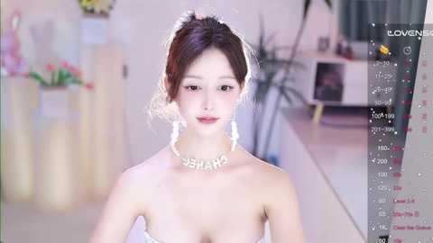 Media: Video of a fair-skinned, East Asian woman with long brown hair, wearing a strapless white dress and a pearl necklace with \"BANANA\" written in large letters, in a softly lit room with a potted plant in the background.