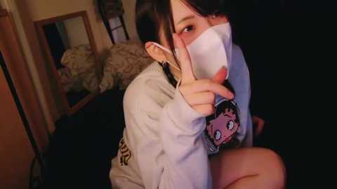 Media: A video of a young woman with short black hair, wearing a face mask, gray hoodie, and a cartoon character shirt, sitting on a bed in a dimly lit room with a mirror and patterned pillow visible in the background.