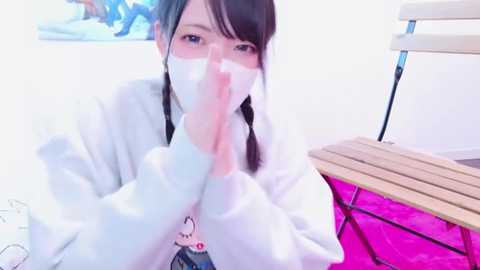 Media: A video of a young Asian woman with long black hair and glasses, wearing a white mask and a white hoodie with a cartoon character, praying indoors.