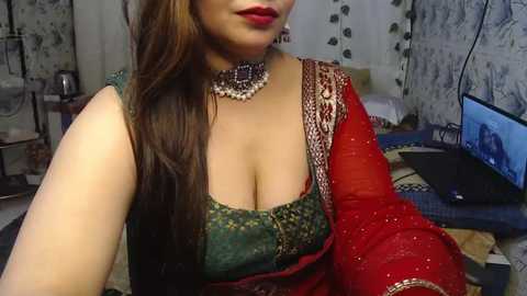 Media: Video of a woman in traditional Indian attire, a green blouse with a deep neckline, red sari, and a beaded choker. Background includes a TV, bed, and decorative wall hangings in a cozy room.