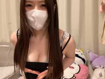 Media: Video of a young Asian woman with long brown hair, wearing a black dress, white mask, and gloves, posing in a room with plush toys, including a Hello Kitty, and a beige couch.