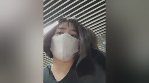 Media: A video shows a woman with dark hair, wearing a white surgical mask and a black jacket, standing in front of a closed, horizontal, silver metal door with slats.