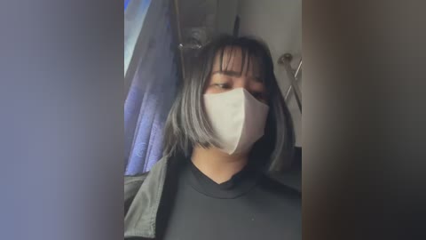 Media: Video of a young woman with medium-length, straight black hair and bangs, wearing a white face mask, black top, and gray jacket. Background includes a blue curtain and a partially visible white wall.