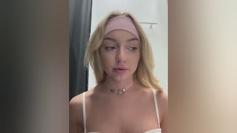 Media: A video of a fair-skinned, blonde woman with a pink headband, wearing a white bralette, standing in front of a mirror. The background is blurry.