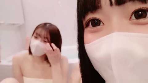 Media: Video of an Asian woman with straight black hair, wearing a white surgical mask, sitting in a white bathtub, looking at the camera.