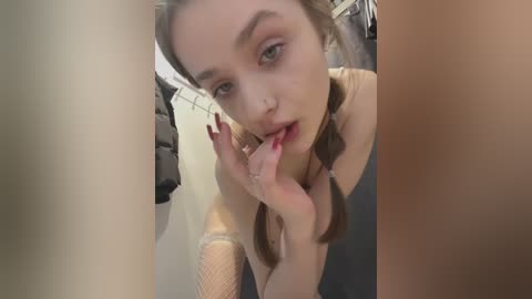 Media: Video of a young Caucasian woman with fair skin, wearing a beige top and fishnet sleeves, licking her finger seductively. She has light brown hair styled in braids. Background shows a dark, possibly industrial setting.